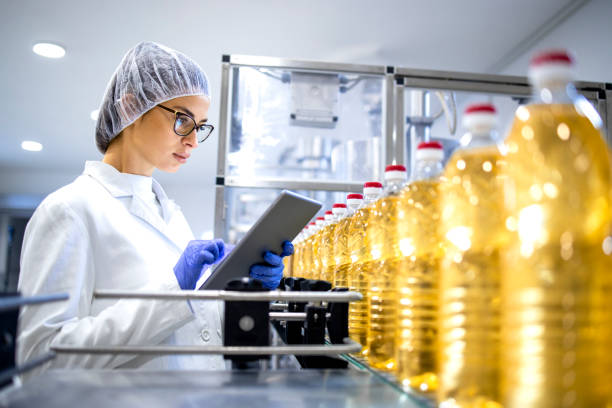 oil worker Food factory worker in sterile white uniform and hairnet using digital tablet while bottled refined oil is being produced on automated conveyor machine.